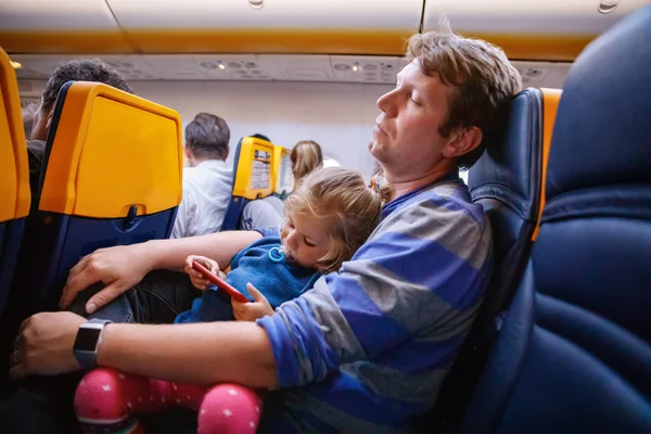 Young father holding his baby toddler daughter during flight on airplane going on vacations. Tired man and cute girl during night flight. child playing with smartphone. Family sitting in plane.