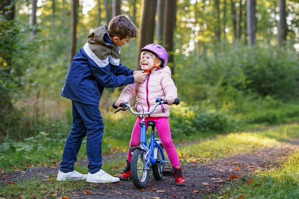 小屁孩 哥哥把小妹妹的自行车头盔戴在了头上 哥哥教快乐的孩子骑自行车和玩自行车 活跃的兄弟姐妹家庭在户外 儿童活动 — 图库照片