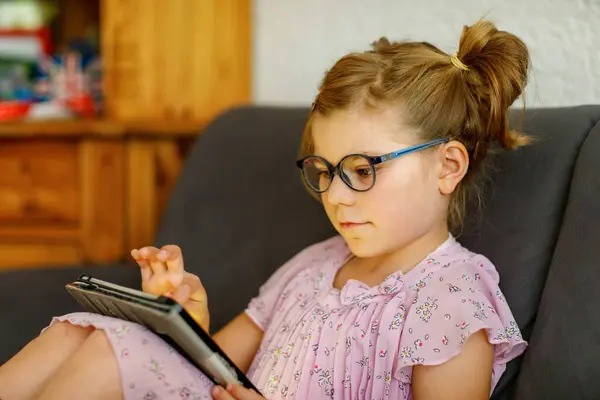 Positive little girl, holding tablet computer in her hands. Preschool child with eyeglasses playing games and learning with different apps