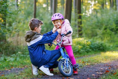 Erkek kardeş, kız kardeş, kız kardeş bisiklet kaskını kafasına taktı. Kardeş mutlu çocuk bisikleti öğretiyor ve bisikletle eğleniyor. Aktif aile fertleri açık havada. Çocuk aktivitesi.