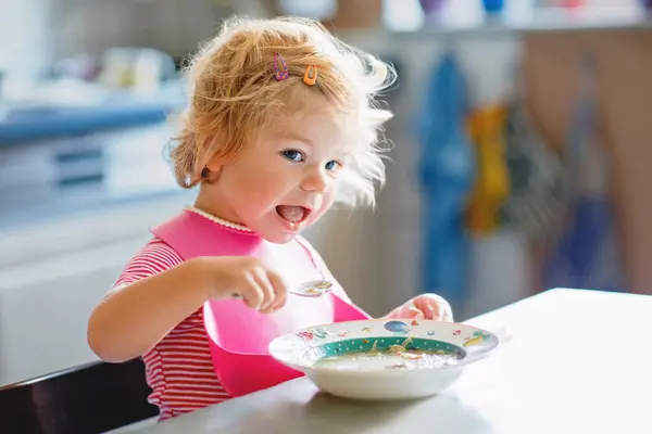 Adorable baby girl eating from spoon vegetable noodle soup. Healthy food, child, feeding and development concept. Cute toddler child with spoon sitting in highchair and learning to eat by itself.