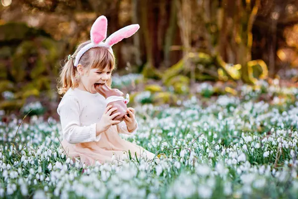 晴れた日に春の森の中でチョコレートフィギュアを食べてイースターバニーの耳を持つ幸せな女の子 たくさんの雪の花を持つかわいい子供 キリスト教の休日の概念 — ストック写真