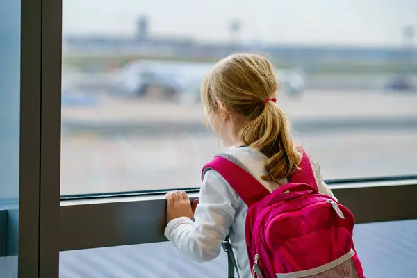 空港の女の子大きな窓で搭乗を待っています かわいい子供は飛行機の背景を背景に窓に立っています 家族の夏休みに向けて出発するのを楽しみにしています — ストック写真