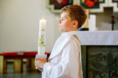 Küçük çocuk ilk kutsal komünyonunu alıyor. Vaftiz mumtutan mutlu çocuk. Katolik curch geleneği. Sunak yakınındaki bir kilisede beyaz geleneksel elbiseli çocuk