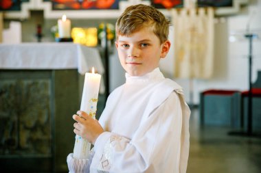 Küçük çocuk ilk kutsal komünyonunu alıyor. Vaftiz mumtutan mutlu çocuk. Katolik curch geleneği. Sunak yakınındaki bir kilisede beyaz geleneksel elbiseli çocuk