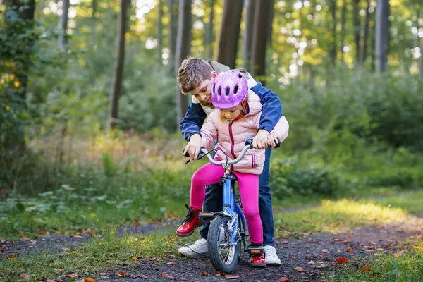 Cute little preschool girl in safety helmet riding bicycle. School kid boy, brother teaching happy healthy sister child cycling and having fun with learning bike. Active siblings family outdoors
