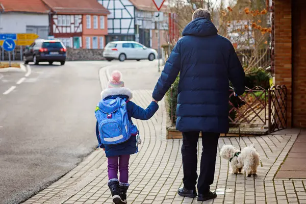 Little girl and middle aged dad on the way to elementary school. Father holding daughter, school child by hand. With little maltese puppy dog. Happy family on the street, back to school