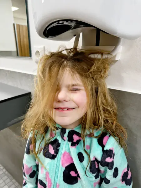 Little smiling girl dries hairs with hairdryer after vistining swimming pool. Happy child with long blond hairs