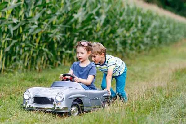 夏の庭で大きな古いおもちゃの車で遊んでいる2人の幸せな子供 中に女の子がいる男の子の運転車 笑いと笑顔の子供たち 子供時代 ライフスタイルの概念 — ストック写真