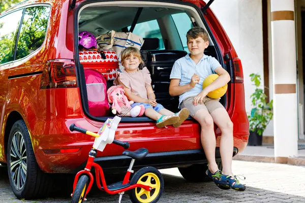 2人の子供 学校の少年と就学前の女の子は夏休みのために両親と一緒に出発する前に車のトランクに座っています 幸せな子供 妹スーツケースとおもちゃが旅に出る — ストック写真