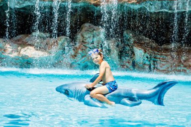 Küçük mutlu çocuk havuza atlıyor ve otel odasında aile tatillerinde eğleniyor. Sağlıklı çocuk şişme köpekbalığı oyuncağıyla suda oynuyor.