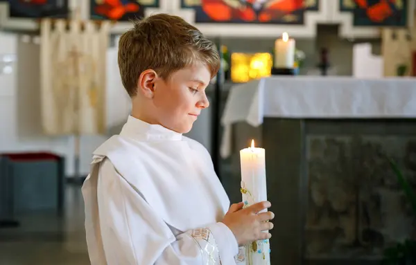 小男孩接受他的第一次圣餐 快乐的孩子拿着洗礼蜡烛 天主教的诅咒的传统 在祭坛附近的教堂里穿着白色传统礼服的孩子 — 图库照片