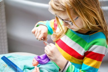 Evde oyuncak bebekle oynayan tatlı küçük bir kız. Gözlüklü çocuk oyuncak bebeğinin saçını kesiyor. Çocuklar için ev içi yaratıcı etkinlikler.