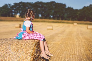 Cute little kid girl in traditional Bavarian costume in wheat field. Happy child with hay bale during Oktoberfest in Munich. Preschool girl play at hay bales during summer harvest time in Germany