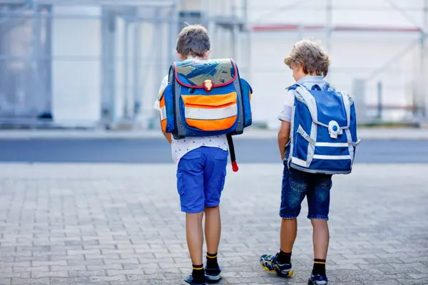 Two little kid boys with backpack or satchel. Schoolkids on the way to school. Healthy adorable children, brothers and best friends outdoors on the street leaving home. Back to school. Happy siblings