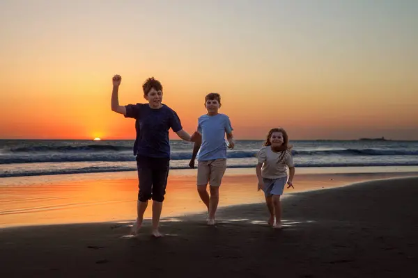 3人の子供が走り 日没時にビーチでジャンプします ハッピーファミリー 2人の学校の少年と1人の幼稚園の少女 一緒に楽しんでいるシンボル ボンディングと家族の休暇 ストック画像