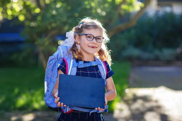 バックパックまたはサテラスと学校の最初の日に大きなスクールバッグで眼鏡をかけた幸せな小さな少女 屋外で健康な愛らしい子供たち チャークデスクを初めて持っている子供 ストック写真