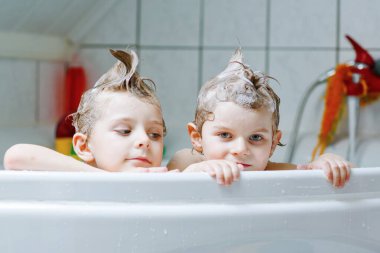 Mutlu kardeşler: İki küçük sağlıklı ikiz çocuk evde küvette banyo yaparak suyla birlikte oynuyorlar. Çocuklar birlikte eğleniyor. Çocuklar saçlarını şampuanla yıkıyor.
