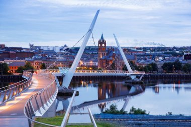 Derry, İrlanda. Kuzey İrlanda 'da, Derry Londonderry' de, Kültür Şehri 'nde şehir merkezinde aydınlatılmış bir barış köprüsü. Gece bulutlu gökyüzü, alacakaranlıkta nehirde yansıyor..