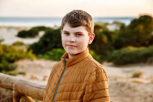 सूर्यास्त पर समुद्र तट पर खड़े खुश किशोर। हैप्पी प्रीटीन सुंदर लड़का कैमरा पर मुस्कुराते हुए। समुद्र में परिवार की छुट्टी पर बच्चे स्टॉक तस्वीर