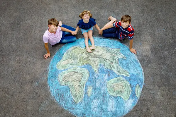 学龄前小女孩和两个小学生 带着绘有彩色粉笔的地球仪画在地上 快乐地球日的概念 为拯救世界 环境和生态而创造儿童 图库照片