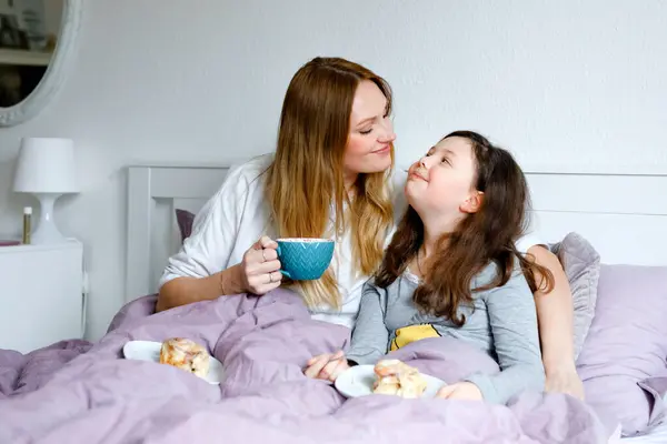 素敵な若いお母さんとかわいい学校の子供の女の子が朝ベッドで一緒に抱き合っています 幸せな家族の若い独身女性と美しい娘朝食を持っています 母の日に食べる ロイヤリティフリーのストック写真