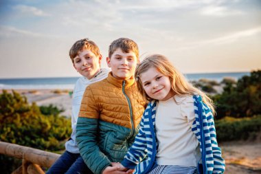 Üç çocuğun portresi, gün batımında sahilde mutlu çocuklar. Mutlu bir aile, iki okul çocuğu ve küçük bir anaokulu kızı. Kardeşler birlikte eğleniyor. Bağ kurmak ve aile tatili.