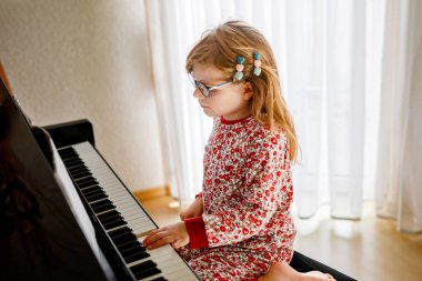 Oturma odasında piyano çalan küçük mutlu kız. Gözlüklü sevimli anaokulu çocuğu müzik aleti çalmayı öğrenirken eğleniyor.