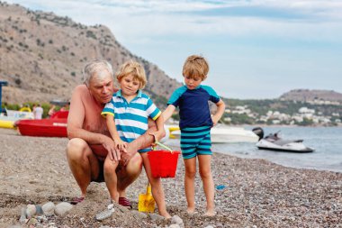 Mutlu büyükbaba ve iki küçük çocuk kumsalda oynuyorlar ve kumdan ve taştan kale yapıyorlar. Güzel bir aile deniz ve okyanusta tatilin tadını çıkarıyor. Büyükbaba ve çocuklar.