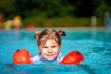 Gün batımında açık havuzda yüzen koruyucu yüzme elbiseli küçük bir anaokulu kızı. Çocuk havuzda yüzmeyi, su sıçratmayı, gülmeyi ve eğlenmeyi öğreniyor. Aile tatilleri