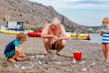 Mutlu büyükbaba ve iki küçük çocuk kumsalda oynuyorlar ve kumdan ve taştan kale yapıyorlar. Güzel bir aile deniz ve okyanusta tatilin tadını çıkarıyor. Büyükbaba ve çocuklar.