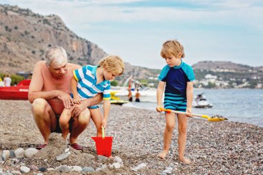 Mutlu büyükbaba ve iki küçük çocuk kumsalda oynuyorlar ve kumdan ve taştan kale yapıyorlar. Güzel bir aile deniz ve okyanusta tatilin tadını çıkarıyor. Büyükbaba ve çocuklar.
