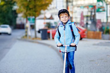 Kasklı aktif okul çocuğu güneşli bir günde şehirde scooterıyla geziyor. Renkli elbiseli mutlu çocuk okula giderken bisiklete biniyor. Çocuklar için güvenli okul yolu..