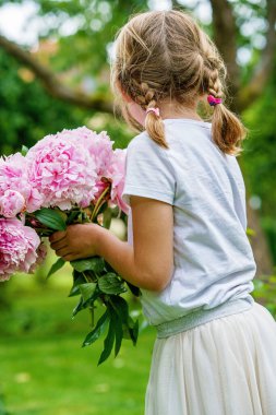 Doğanın arka planında çiçek açmış, el ele tutuşan, koklayan ve bir buket pembe şakayık çiçeğinin tadını çıkaran, mutlu, küçük beyaz bir kız çocuğunun portresi.
