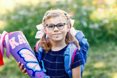 Gözlüklü ve mavi bir sırt çantası olan, denizci üniforması giyen küçük bir kız, Schultuete adında renkli bir okul konisi ve sırt çantası taşıyor. Mutlu çocuk gülümsüyor, okulun ilk günü için hazır.