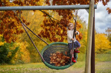 Sonbaharda salıncakta sallanan mutlu kız çocuğu. Sonbaharda parkta oynayan, sallanan ve eğlenen küçük bir çocuk. Arka planda sonbahar ağaçları var. Okulun arka bahçesi