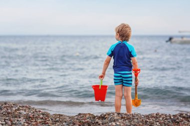 Yalnız okyanus sahilinde duran sevimli sarışın çocuk. Çocuk kova ve kürekle oynuyor ve ufuk çizgisine bakıyor. Tatiller, yaz tatili, seyahat konsepti. Yaz tatillerinde deniz kenarında