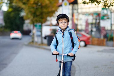Kasklı aktif okul çocuğu güneşli bir günde şehirde scooterıyla geziyor. Renkli elbiseli mutlu çocuk okula giderken bisiklete biniyor. Çocuklar için güvenli okul yolu..