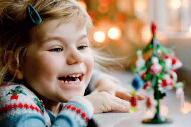 Küçük kız pencerenin kenarında oturuyor ve küçük cam Noel ağacını küçük Noel oyuncaklarıyla süslüyor. Mutlu sağlıklı çocuk geleneksel aile bayramını kutluyor. Sevimli bebek