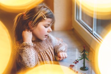 Küçük anaokulu kızı pencerenin yanında oturuyor ve küçük cam Noel ağacını küçük Noel oyuncaklarıyla süslüyor. Mutlu sağlıklı çocuk geleneksel aile tatilini kutluyor.