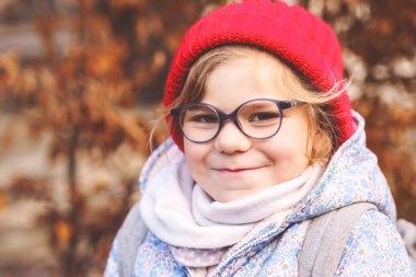 Açık havada gözlüklü ve kırmızı şapkalı sevimli bir anaokulu kızının portresi. Yeni mavi gözlük takan mutlu komik çocuk. Şehirde sonbahar günü