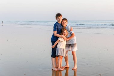 Üç çocuk, gün batımında okyanus sahilinde mutlu kardeşler. Mutlu bir aile, iki okul çocuğu ve küçük bir anaokulu kızı. Kardeşler eğleniyor, İspanya, Costa del Sol