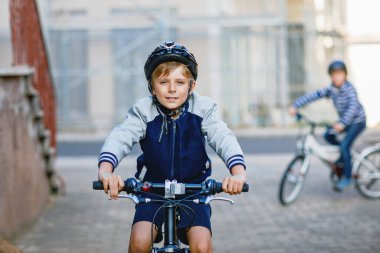 Kasklı iki okul çocuğu şehirde sırt çantalı bisikletle dolaşıyorlar. Renkli giysiler içinde okula bisikletle giden mutlu çocuklar. Çocuklar için güvenli okul yolu..