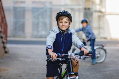 Kasklı iki okul çocuğu şehirde sırt çantalı bisikletle dolaşıyorlar. Renkli giysiler içinde okula bisikletle giden mutlu çocuklar. Çocuklar için güvenli okul yolu..