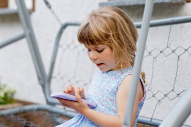 Akıllı telefonla oynayan tatlı küçük kız sağlıklı bebek cep telefonuna parmaklarıyla dokunuyor, çizgi film izliyor ve eğitim uygulamalarıyla eğleniyor. Bahçedeki çocuk..