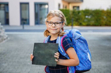 Happy little kid girl with eye glasses with backpack or satchel on the first day of school. Healthy adorable child outdoors. Child holding chalk desk with copyspace for text. Back to school. clipart