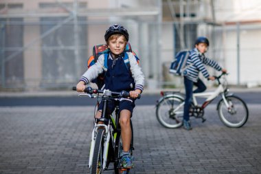 Kasklı iki okul çocuğu şehirde sırt çantalı bisikletle dolaşıyorlar. Renkli giysiler içinde okula bisikletle giden mutlu çocuklar. Çocuklar için güvenli okul yolu..