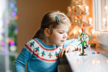 Küçük kız pencerenin kenarında oturuyor ve küçük cam Noel ağacını küçük Noel oyuncaklarıyla süslüyor. Mutlu sağlıklı çocuk geleneksel aile bayramını kutluyor. Sevimli bebek