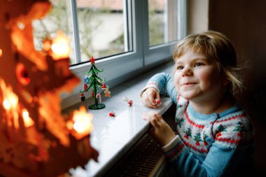 Küçük kız pencerenin kenarında oturuyor ve küçük cam Noel ağacını küçük Noel oyuncaklarıyla süslüyor. Mutlu sağlıklı çocuk geleneksel aile bayramını kutluyor. Sevimli bebek