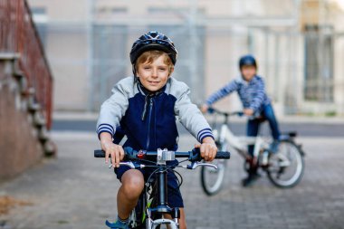 Kasklı iki okul çocuğu şehirde sırt çantalı bisikletle dolaşıyorlar. Renkli giysiler içinde okula bisikletle giden mutlu çocuklar. Çocuklar için güvenli okul yolu..